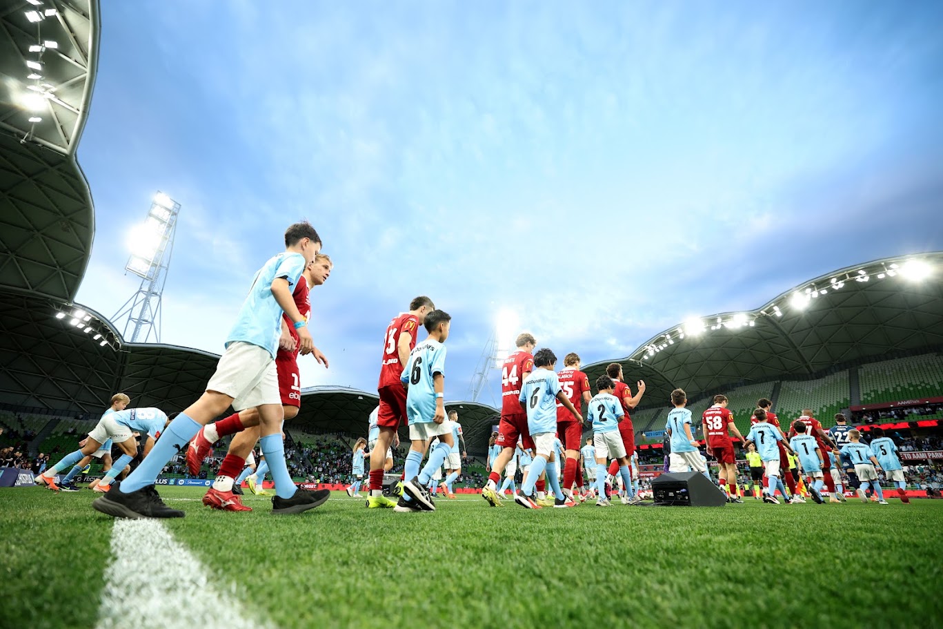 Adelaide United players and child mascots walking out onto the pitch beneath the stadium roof