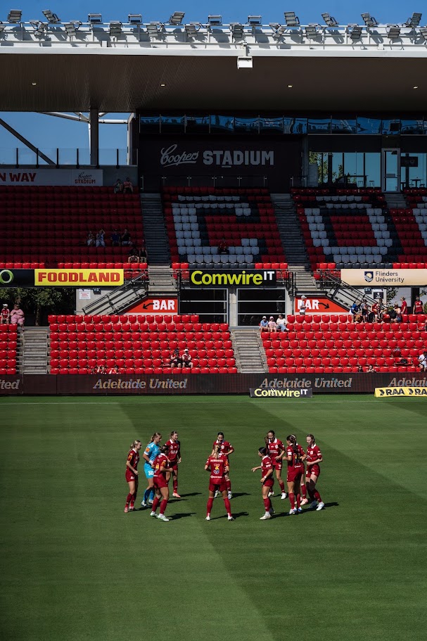 The Adelaide United women's team in a huddle on the pitch at Coopers Stadium on a sunny day